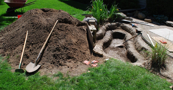 A backyard with a small, shelved hole on the side. Next to the hole is a shovel, a large pile of dirt, and a wheelbarrow.