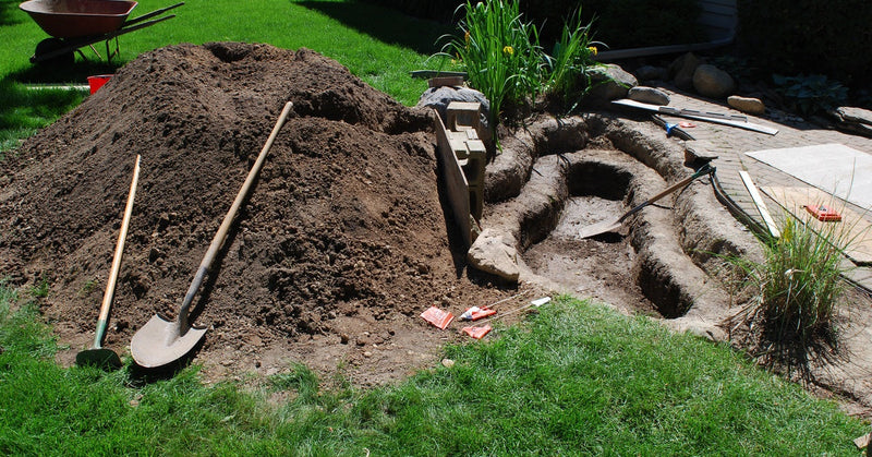 A backyard with a small, shelved hole on the side. Next to the hole is a shovel, a large pile of dirt, and a wheelbarrow.